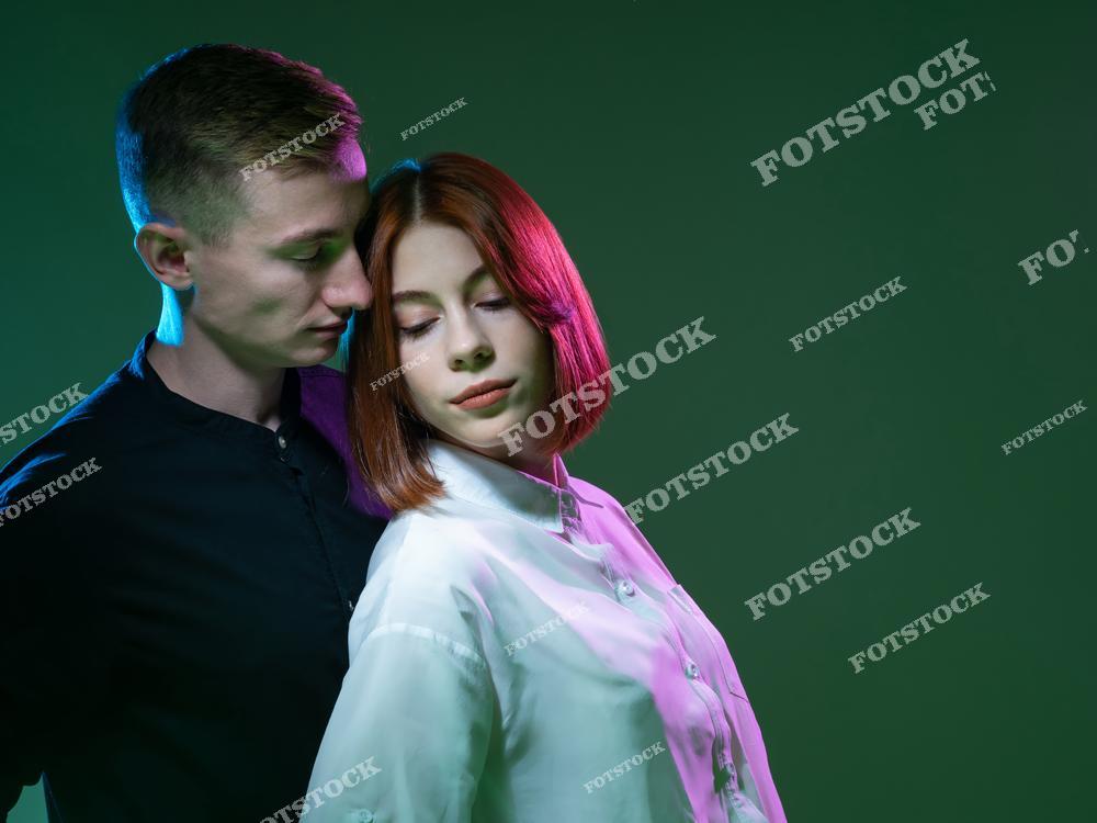 На фотографии изображены мужчина и женщина, стоящие рядом в мягком, интимном объятии. Мужчина нежно смотрит на девушку, а она опускает взгляд, создавая атмосферу доверия и романтики. Цветное освещение в зеленых и розовых тонах добавляет снимку художественный и современный стиль.