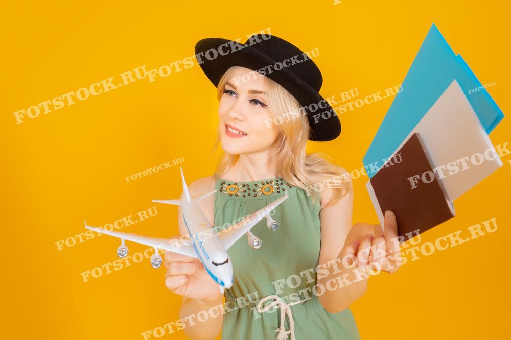 На фотографии изображена молодая женщина в черной шляпе, держащая модель самолета в одной руке и документы для путешествия в другой. Она улыбается, выражая радостное ожидание предстоящего приключения. Яркий желтый фон подчеркивает позитивное настроение и создает атмосферу легкости и радости.