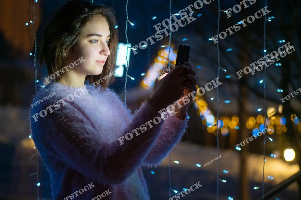 На фотографии изображена женщина, читающая сообщение на телефоне у окна ночью. Она получает поздравление с Рождеством или Новым годом от родственников. Сцена создает атмосферу тепла и связи с близкими в праздничный вечер.
