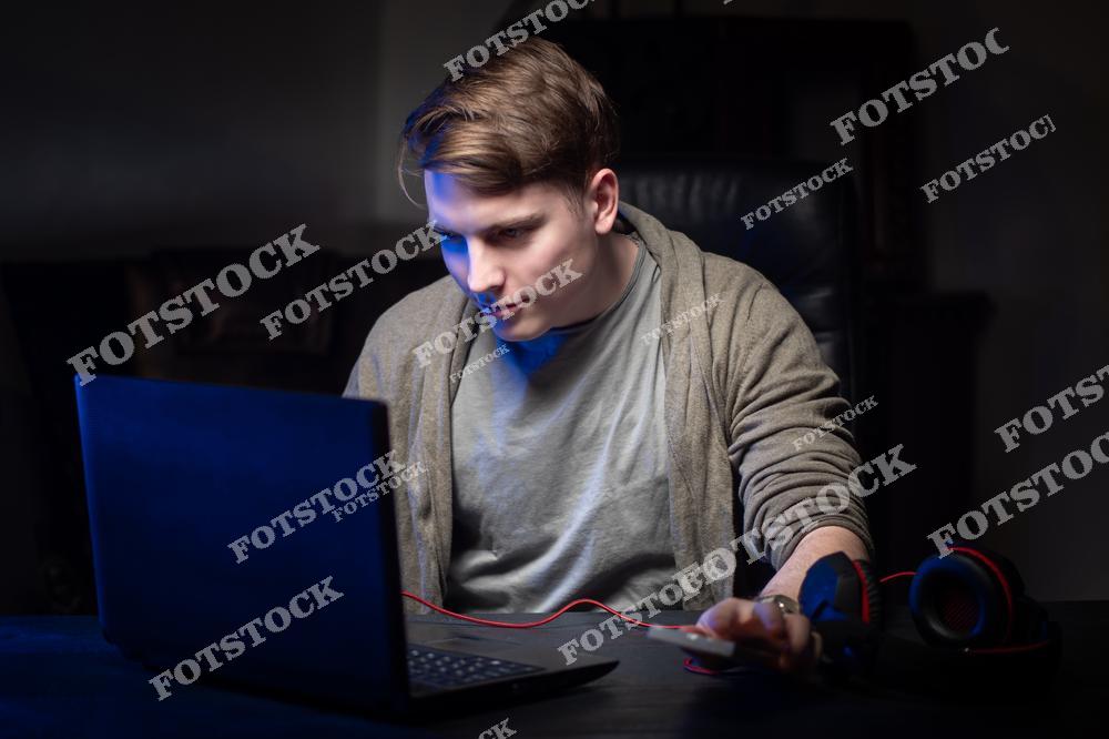 На фотографии изображен молодой человек, сосредоточенно работающий за ноутбуком. Внимание акцентировано на его лице, освещенном экраном, что создает атмосферу уединенности. На столе также видны наушники, подчеркивающие современный стиль работы или отдыха.