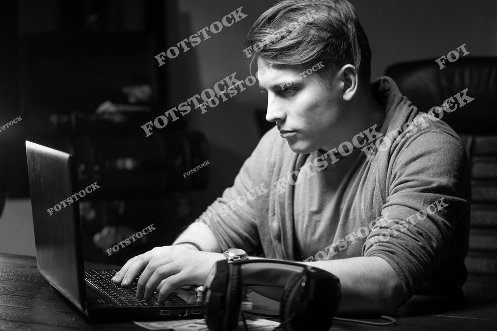 Черно-белая фотография показывает молодого человека, который сосредоточенно работает за ноутбуком. Наушники лежат рядом, создавая атмосферу погружения в процесс. Возможно, это работа фрилансера, программирование или учеба ночью. Обстановка подчеркивает напряженную концентрацию и трудолюбие.