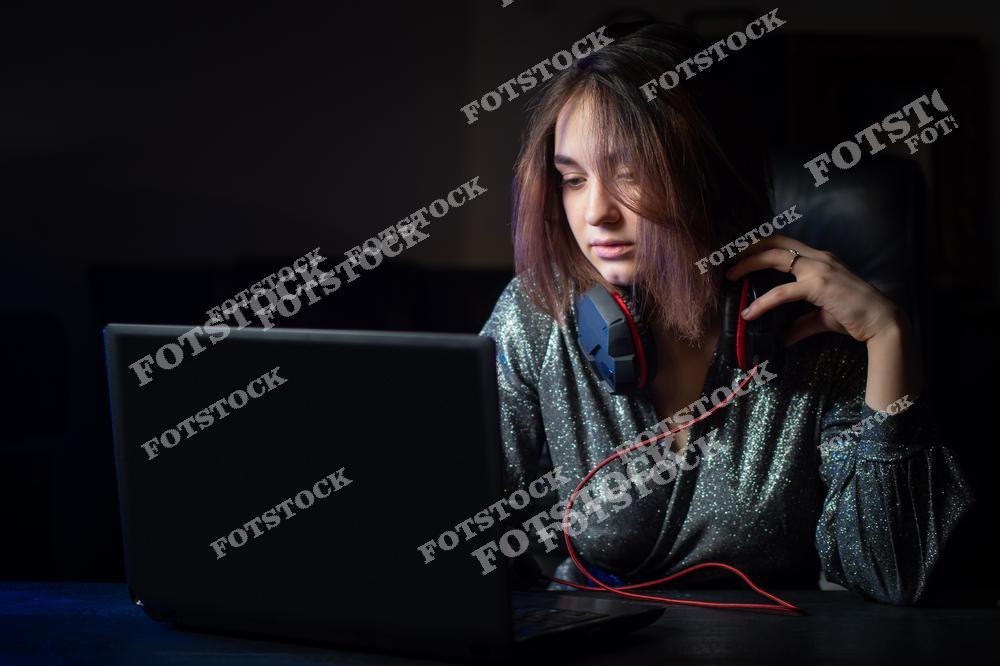 На фотографии изображена молодая женщина в блестящей одежде, сидящая за ноутбуком. Она слегка касается своих наушников, создавая атмосферу спокойной сосредоточенности. Мягкое освещение подчеркивает вечернюю рабочую обстановку и стильный образ.