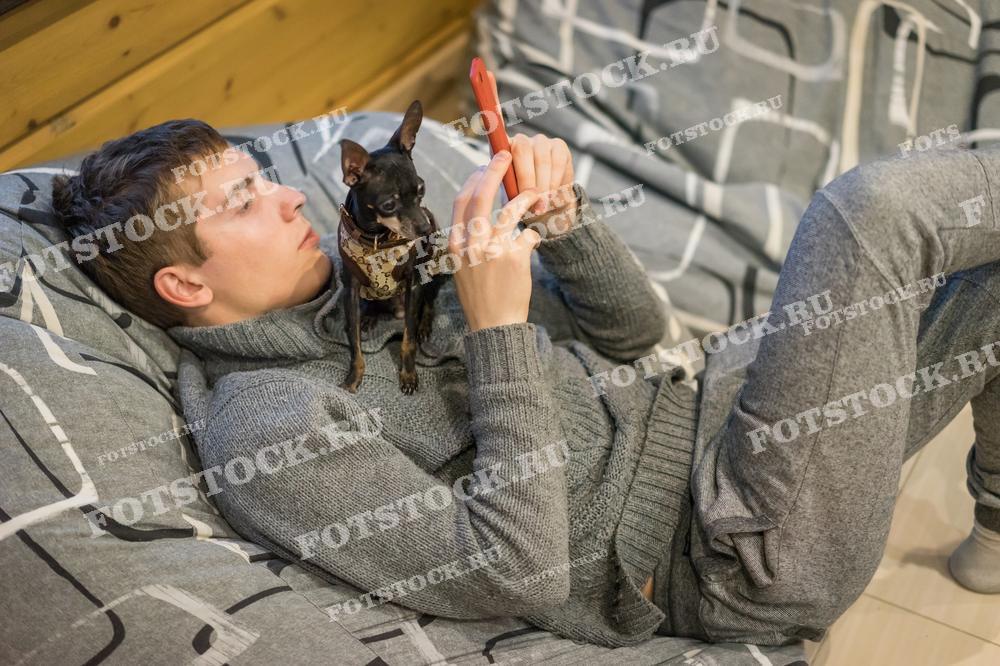 На фотографии изображен молодой человек, лежащий на диване в уютной домашней обстановке. Рядом с ним сидит маленькая собака, а в руках у него смартфон. Мужчина одет в теплую домашнюю одежду. Сцена отражает современный образ жизни, сочетающий домашний уют с использованием цифровых технологий.