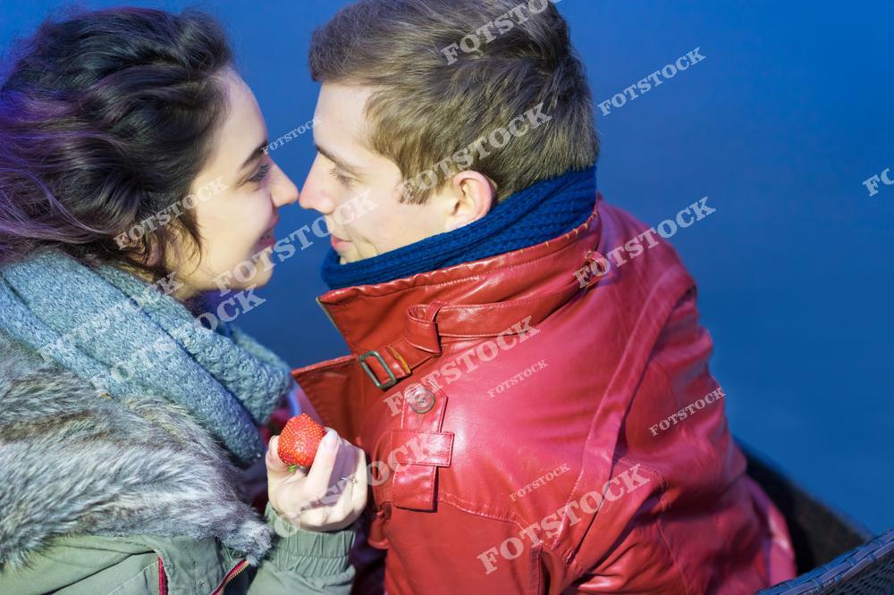 На фотографии изображены мужчина и женщина в теплой одежде, сидящие рядом на фоне синего неба или воды. Девушка держит в руке клубнику, пара смотрит друг на друга с любовью и нежностью. Снимок передает атмосферу романтики и тепла в прохладное время года.