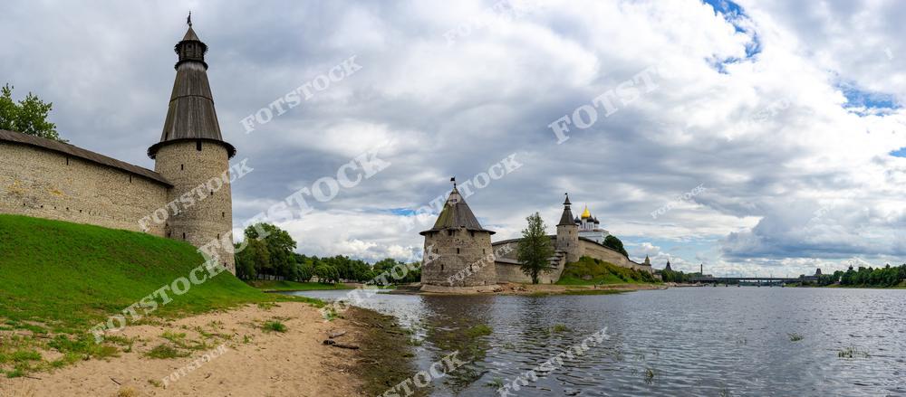 На фотографии представлена панорама Псковского кремля, включающая башни, стены и собор с золотыми куполами. Берег реки с песком и зеленью подчёркивает гармонию архитектуры и природы. Эта сцена передаёт величие и историческое значение Пскова.
