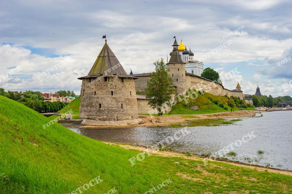 На фотографии изображён Псковский кремль с его мощными башнями и собором с золотыми куполами, расположенный на берегу реки. Зелёные склоны и яркое небо подчёркивают красоту и гармонию этой исторической достопримечательности. Кремль является символом древней Руси и уникальной архитектуры.