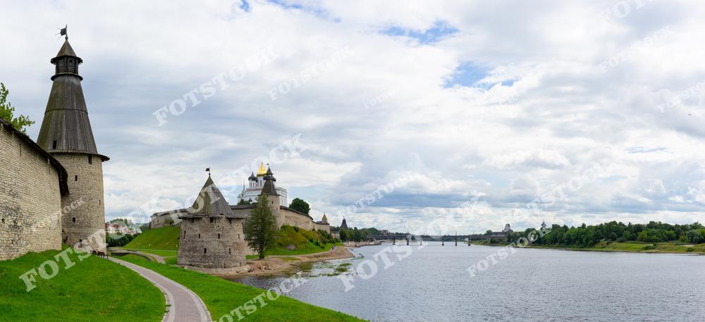 На фотографии изображена панорама Псковского Крома с массивными стенами, башнями и Троицким собором с золотыми куполами на берегу реки Великой. Ясное небо и зелёные склоны добавляют композиции спокойствие и гармонию. Этот пейзаж отражает богатую историю Пскова и его архитектурное наследие.
