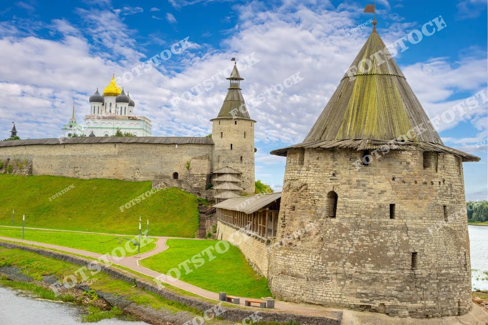На фотографии изображён Псковский Кром, включающий массивные башни, крепостные стены и величественный Троицкий собор с золотыми куполами. Зелёные склоны, голубое небо и архитектурные элементы подчёркивают гармонию природной и исторической красоты. Этот вид представляет одно из главных наследий древней Руси.