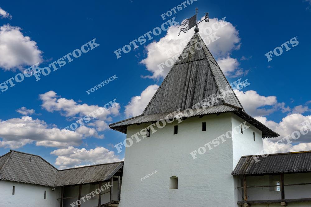 На фотографии изображена крепостная башня Псковского Крома с деревянной крышей и белоснежными стенами на фоне голубого неба с облаками. Древнерусская архитектура подчеркивает историческое значение этого места. Эта башня — часть защитного сооружения и культурное наследие города.