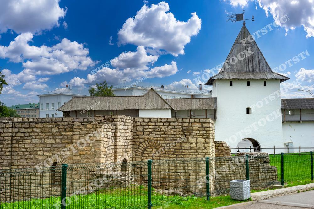 Фотография изображает участок древних каменных стен Псковского кремля с одной из его башен. На фоне чистого голубого неба башня с флюгером подчеркивает уникальность и величие архитектурного ансамбля. Вид огороженных стен создает ощущение связи с историей и культурным прошлым.