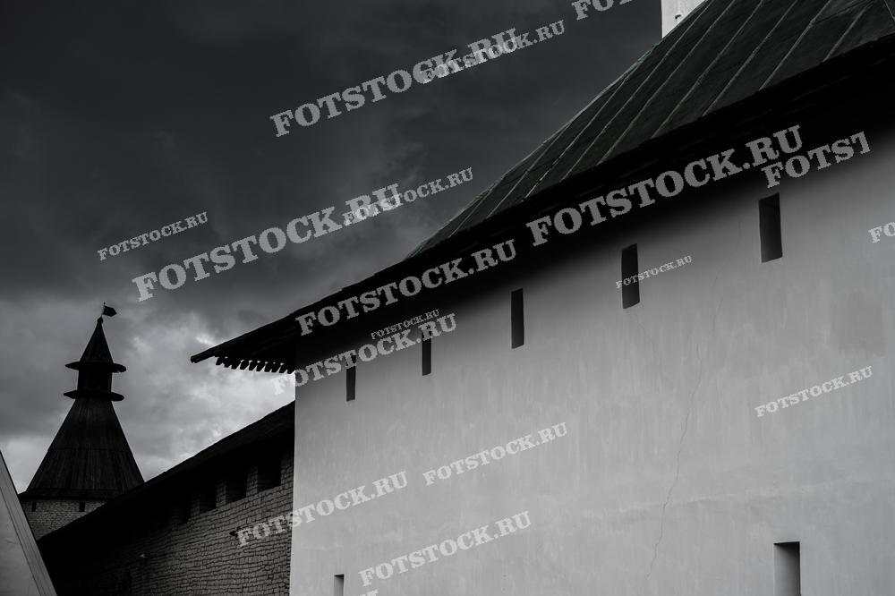 На фотографии изображены стены и башни Псковского Кремля на фоне тёмного грозового неба. Драматическое освещение подчеркивает величие и историческую значимость этого средневекового укрепления.