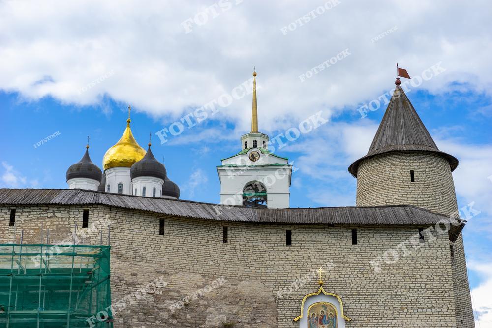 Древняя крепость под ясным небом. Каменные стены и защитная башня с деревянным флагом, в центре виднеются золотые купола православной церкви с зелёным куполом часовни. Башня и стены окружены зелёной травой. Псков, Россия