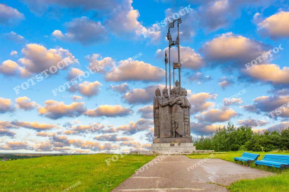 На фотографии изображён памятник Александру Невскому в Пскове. Скульптура выполнена в монументальном стиле и установлена на зелёной лужайке с дорожкой и скамейками. Фон из мягких облаков и голубого неба подчёркивает величественность памятника.