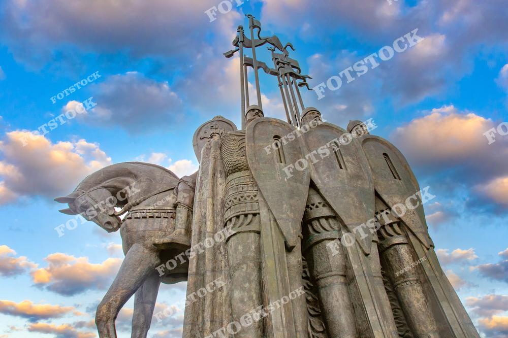 На фотографии изображён крупный план памятника Александру Невскому в Пскове. Скульптура включает фигуры рыцарей с щитами и коня, выполненные в монументальном стиле. Фон из облаков подчёркивает величие композиции и историческую значимость памятника.