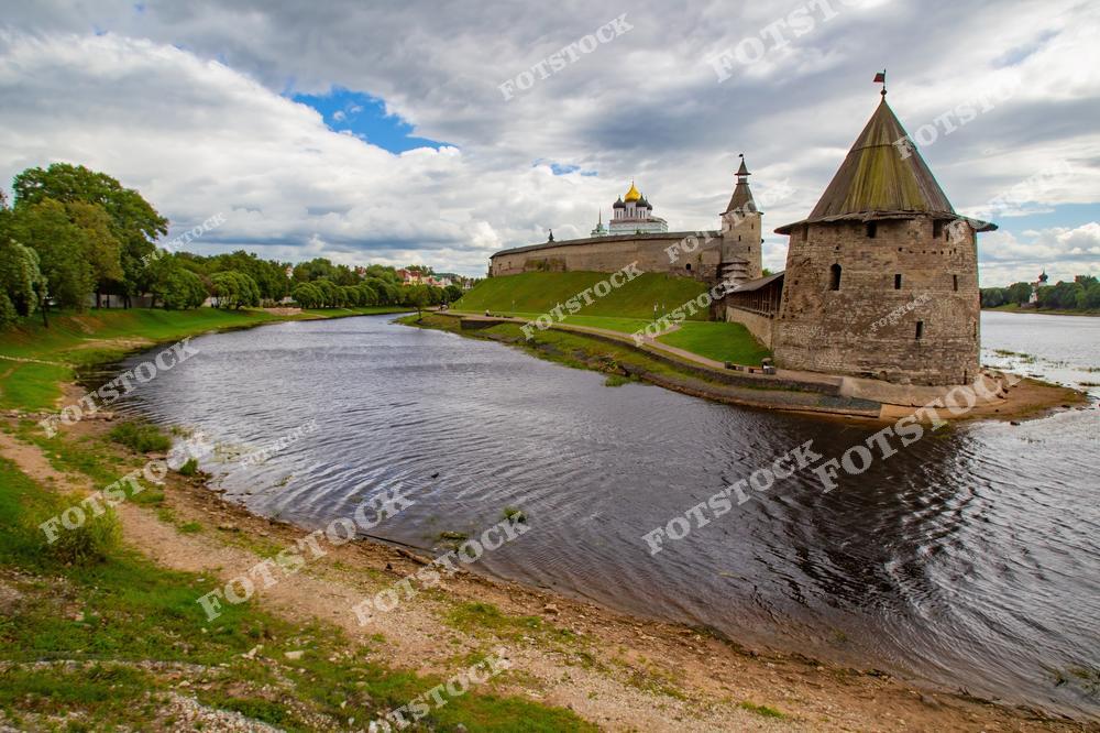 На фотографии изображён Псковский Кремль, окружённый рекой Великой. В центре видны древние крепостные башни и купола собора, возвышающиеся над зелёными холмами. Пейзаж передаёт величие архитектуры и спокойствие природной среды.