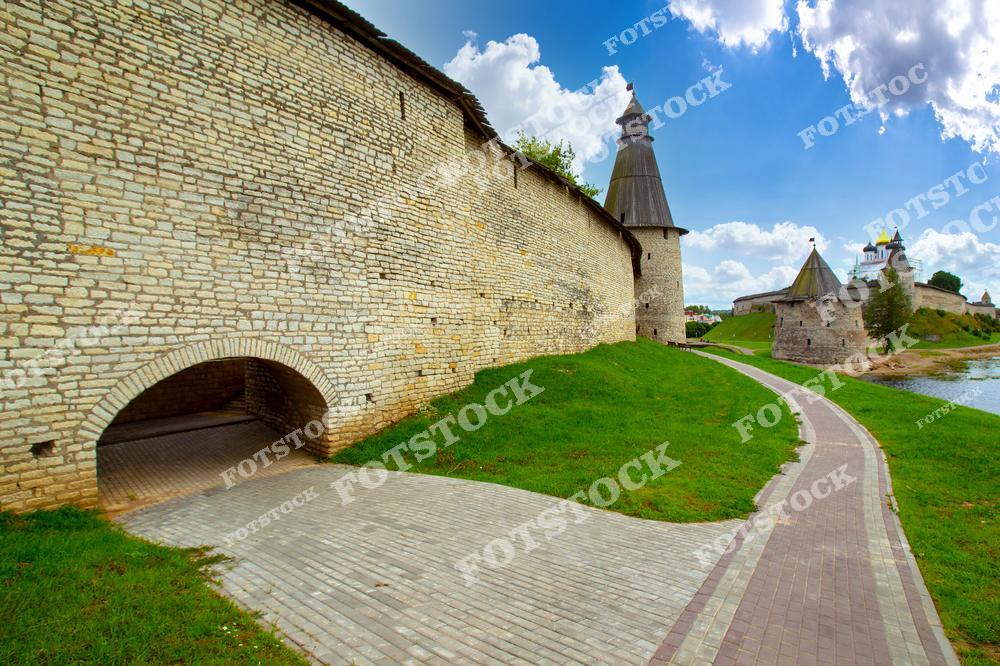 На фотографии изображён Псковский Кремль с его массивными крепостными стенами и башнями. Пешеходная дорожка ведёт вдоль зелёного газона к историческим сооружениям, расположенным на берегу реки. Композиция подчёркивает уникальность средневековой архитектуры.