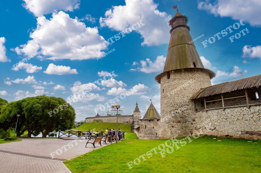 На фотографии изображена часть Псковского Кремля с массивной башней и крепостными стенами. На переднем плане видна прогулочная зона с туристами, расположенная рядом с зелёной лужайкой. Голубое небо с облаками создаёт атмосферу спокойствия и величия.