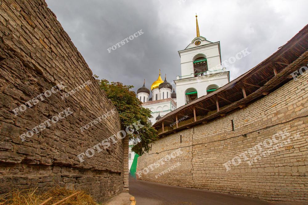 На фотографии изображён Троицкий собор в Пскове, окружённый массивными крепостными стенами. Золотые купола собора выделяются на фоне тёмного грозового неба, создавая драматичный контраст. Композиция подчёркивает величие древнерусской архитектуры и её историческую значимость.
