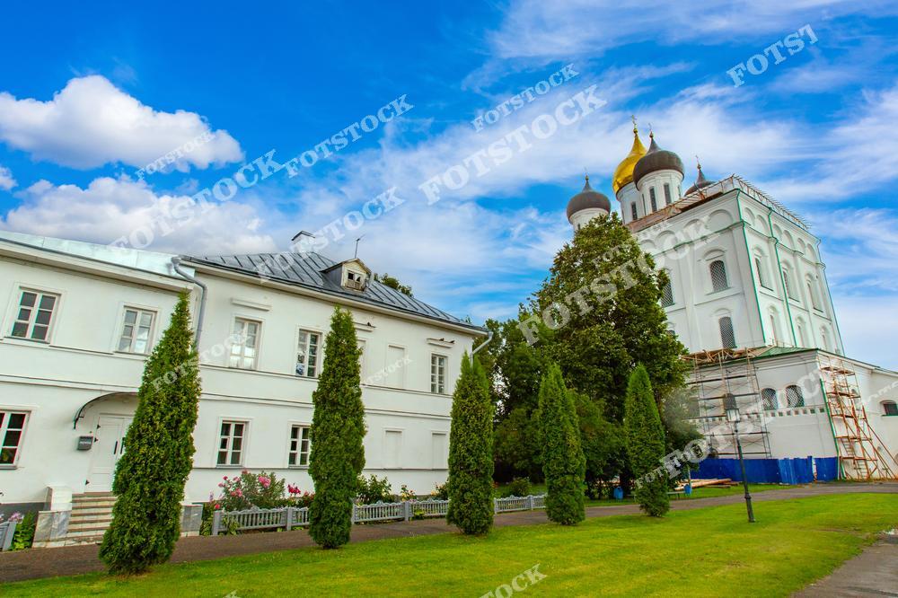 На фотографии изображён Троицкий собор с золотыми куполами, а также прилегающие здания на территории Псковского Кремля. Зелёные насаждения и голубое небо создают гармоничное обрамление архитектурного ансамбля. Пространство передаёт атмосферу истории и духовности.