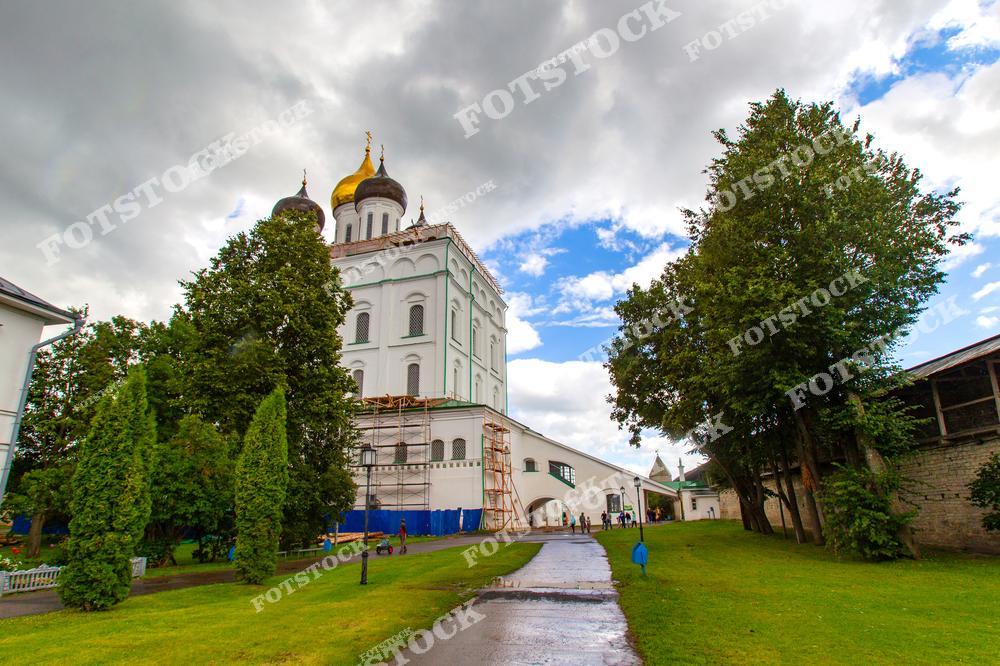 На фотографии изображён Троицкий собор в Пскове с золотыми куполами, окружённый деревьями и зелёной лужайкой. Пешеходная дорожка ведёт к храму, подчёркивая гармонию архитектуры и природы. Контрастное небо создаёт атмосферу величия и духовности.