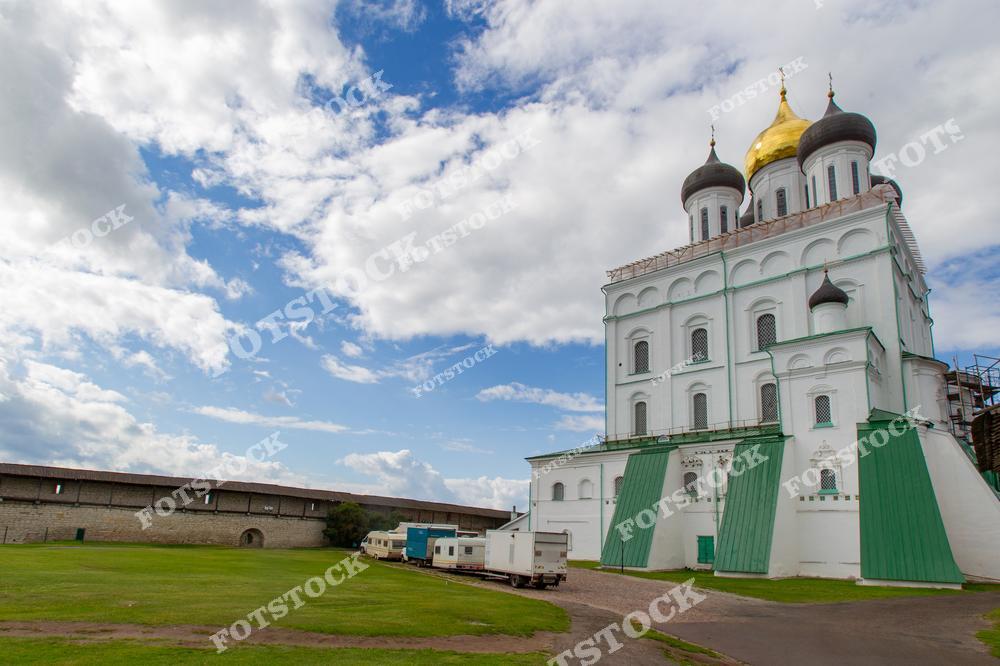 На фотографии изображён Троицкий собор в Пскове с золотыми куполами, окружающая территория украшена зелёной лужайкой. Белокаменные стены и крепостные сооружения дополняют ансамбль. Голубое небо с облаками подчёркивает красоту исторической архитектуры.