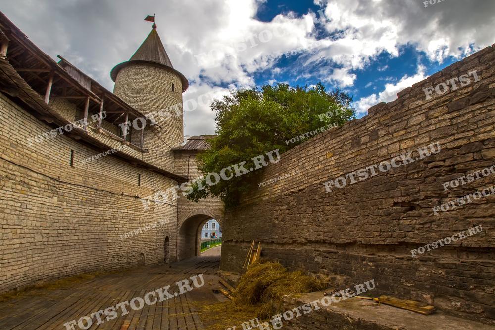 На фотографии изображена древняя каменная крепость под облачным небом. Это одна из башен Псковского Кремля и его крепостные стены. Данное место является исторической достопримечательностью Псковщины и привлекает много туристов.