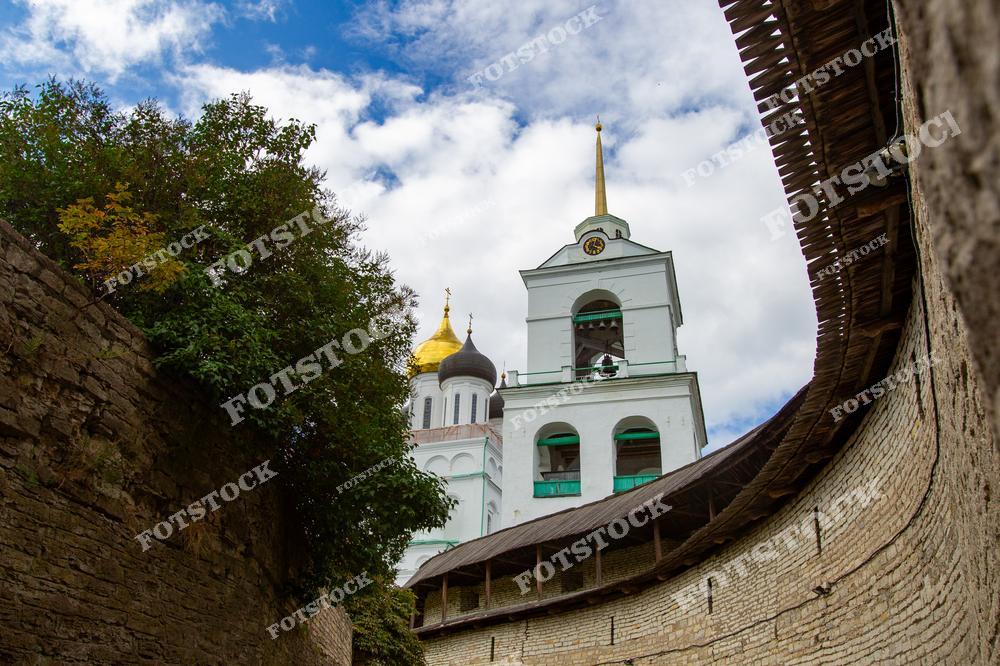 Уникальный ракурс, захватывающий вид на колокольню и Троицкий собор Псковского Кремля. Высокие крепостные стены обрамляют золотые купола и шпиль, создавая атмосферу древности и величия. Листва дерева добавляет природную мягкость к суровости камня.