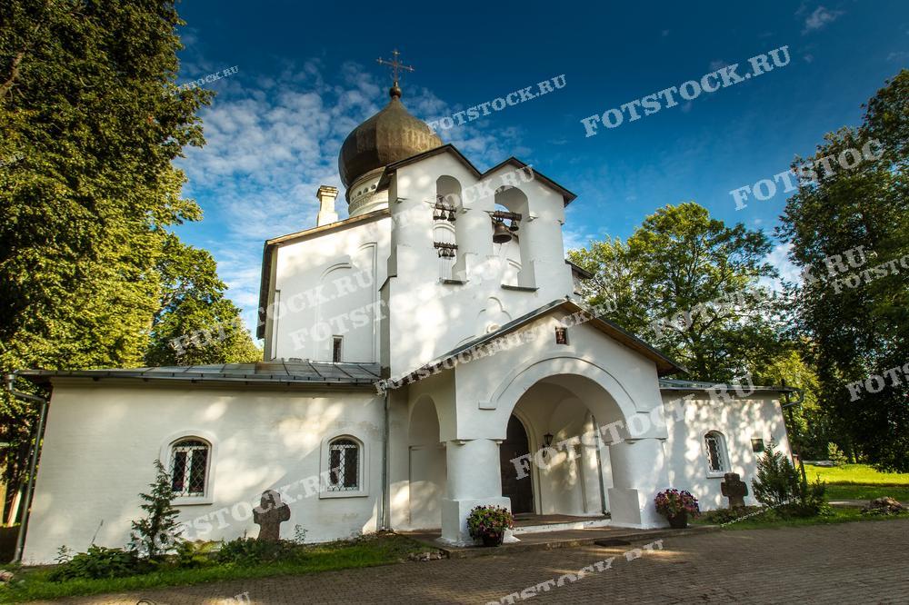 На фотографии изображена белокаменная церковь в Гдове, окруженная зелеными деревьями парка. Православный храм выделяется на фоне голубого неба, создавая атмосферу спокойствия и умиротворения.