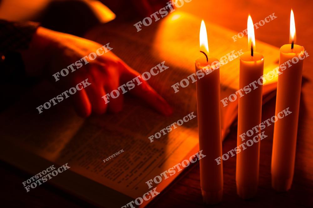 На фото три горящие свечи, стоящие рядом с открытой книгой, которую освещает их теплое пламя. Рука человека указывает на текст, подчеркивая момент чтения или размышления в спокойной и духовной атмосфере.