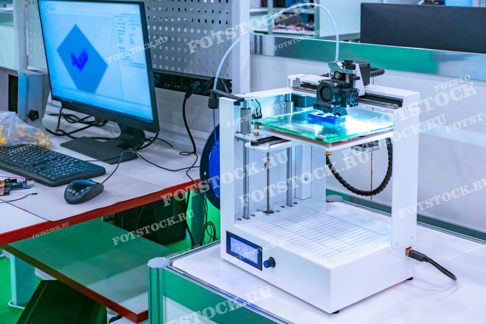 Фотография показывает 3D-принтер на рабочем столе в лабораторной обстановке. Рядом с принтером находится компьютер с программным обеспечением для 3D-моделирования. Изображение иллюстрирует процесс создания объектов с помощью аддитивных технологий.