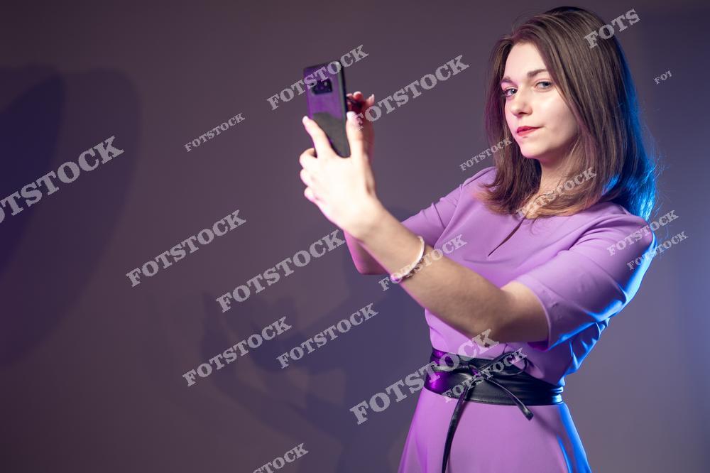 На фото изображена молодая деловая женщина, держащая в руках смартфон. Она смотрит в камеру, демонстрируя уверенность и современный стиль работы с мобильными гаджетами в бизнес-среде.
