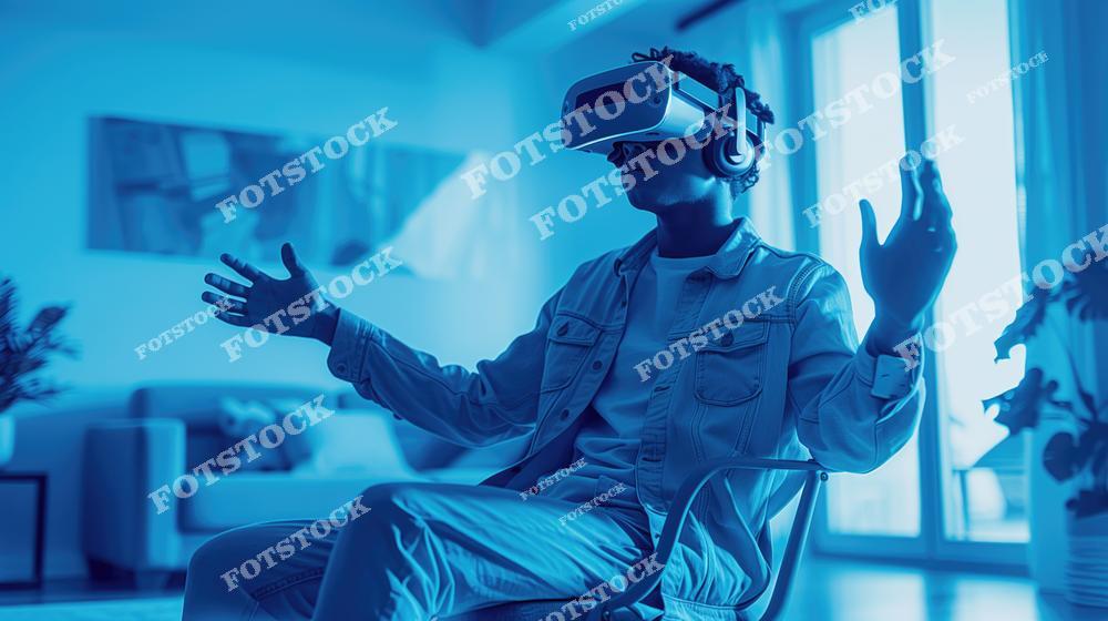 На фотографии изображен мужчина, сидящий в комнате и использующий VR-гарнитуру. Голубое освещение подчеркивает технологичность и атмосферу полного погружения. Пространство комнаты создает ощущение уюта и современности.