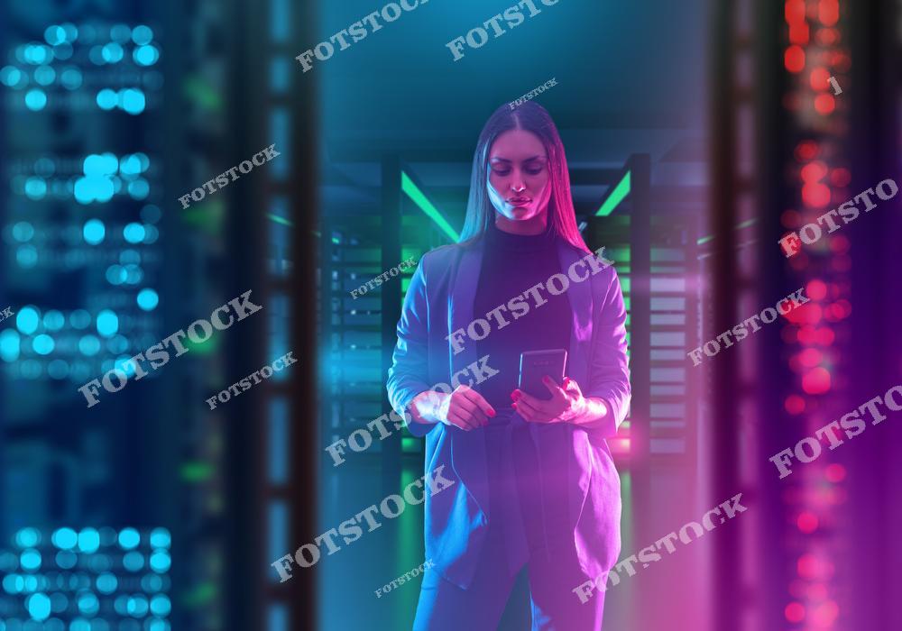 На изображении молодая женщина в строгом костюме, работающая с планшетом на фоне серверной комнаты с яркими огнями и технологической атмосферой. Фотография символизирует управление данными, работу в IT-индустрии и развитие современных технологий.