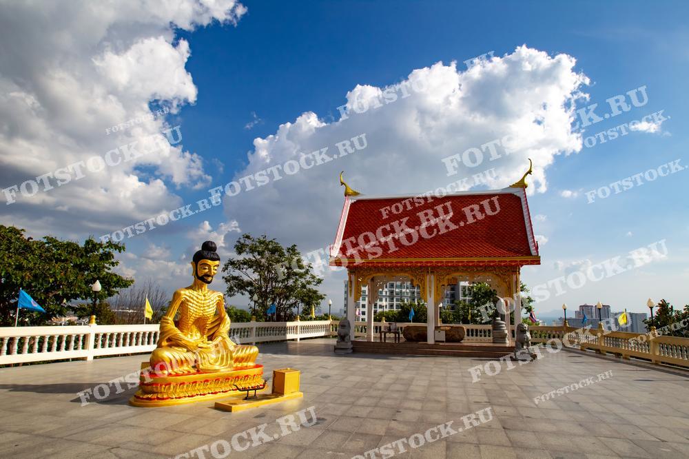 Фотография показывает золотую статую Будды, сидящую на открытой террасе рядом с традиционным тайским храмом с красной крышей. На фоне виден городской пейзаж Паттайи и голубое небо с белыми облаками. Изображение передает атмосферу спокойствия и духовности, типичную для храмов Таиланда.