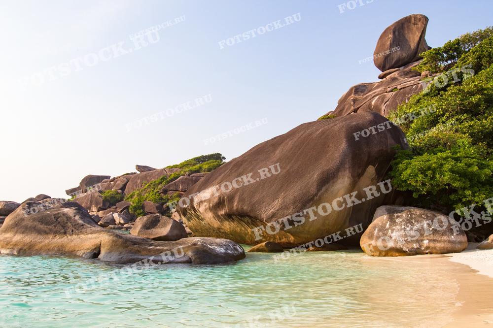 На фотографии изображён красивый пляж с белым песком и бирюзовой водой на Симиланских островах. Массивные гранитные скалы и зелёные растения окружают берег, создавая идиллический тропический пейзаж.
