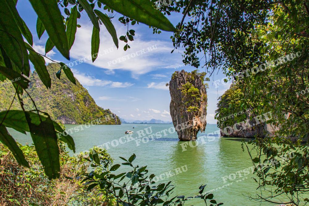 На фото изображён остров Джеймса Бонда в заливе Phang Nga, окружённый бирюзовыми водами и тропической зеленью. Снимок сделан через густую растительность, что создаёт естественную рамку для скалы, возвышающейся из воды. Ясное небо и спокойная вода дополняют идиллический пейзаж.
