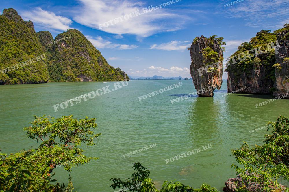 На изображении живописный вид на остров Джеймса Бонда в заливе Phang Nga, Таиланд. Скала возвышается из воды, окружённая другими зелёными холмами и скалами. Бирюзовая вода и ясное небо создают впечатление спокойствия и гармонии с природой.