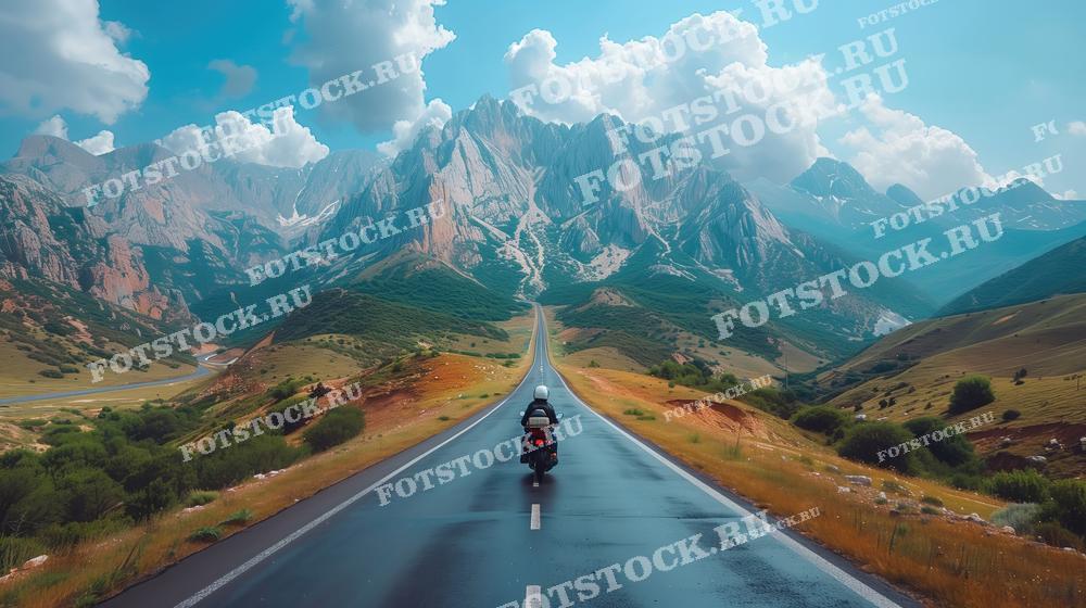 Фотография мотоциклиста, едущего по живописной горной дороге. Величественные горы и облака создают атмосферу свободы и приключения, подчеркивая красоту природного ландшафта и дух путешествия.
