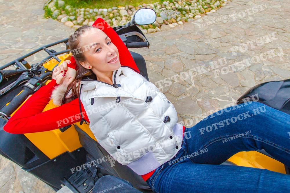 На фотографии молодая женщина в белой жилетке и красном свитере расслабленно лежит на квадроцикле на фоне каменной дороги. Эта сцена демонстрирует отдых и удовольствие от активного отдыха на свежем воздухе.