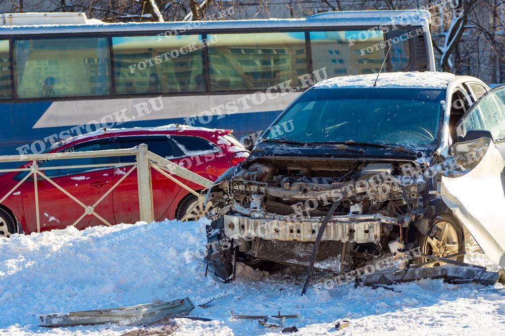 На фотографии изображен сильно поврежденный автомобиль на заснеженной улице. Автомобиль стоит рядом с автобусом и красной машиной на фоне городского пейзажа. Это иллюстрация последствий дорожной аварии в зимних условиях.