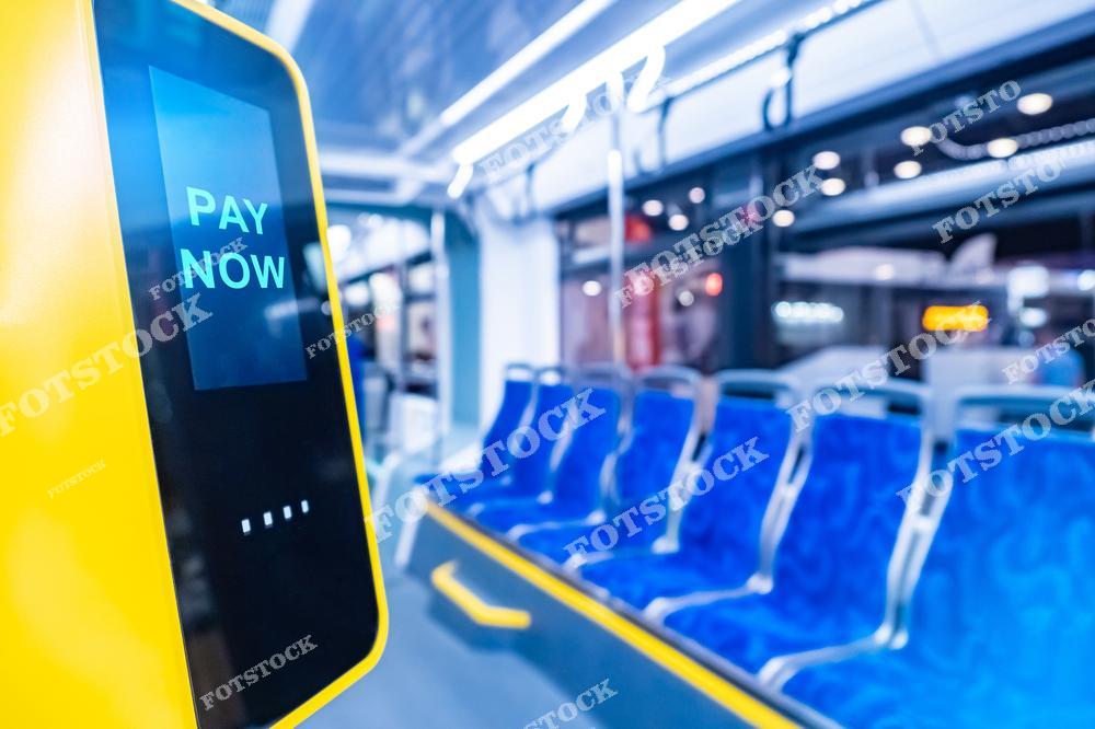 На фотографии изображен терминал оплаты с надписью "Pay Now" в салоне автобуса. Сиденья синего цвета и яркий свет создают современную атмосферу общественного транспорта. Изображение подчеркивает удобство и технологичность системы оплаты.