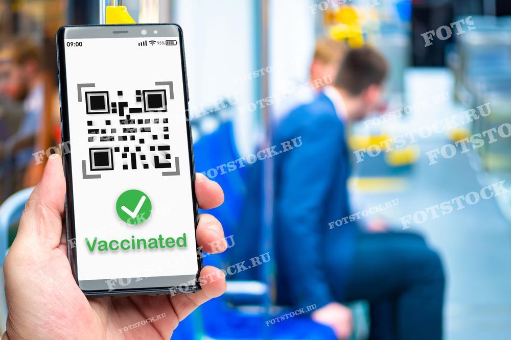 На фотографии изображена рука, держащая смартфон с QR-кодом и отметкой о вакцинации. На фоне человек сидит в общественном транспорте, что символизирует использование цифровых технологий для подтверждения статуса вакцинации.