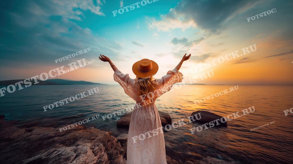 На фотографии изображена женщина в легком платье и шляпе, стоящая на каменистом берегу и поднявшая руки к закатному небу. Она смотрит на море, которое окрасилось в теплые оттенки заката. Пейзаж создает атмосферу свободы, спокойствия и наслаждения природой. Эта фотография может быть использована для иллюстрации материалов о путешествиях, медитации, отдыхе на природе и романтике, а также в рекламных кампаниях туристических агентств и курортов.