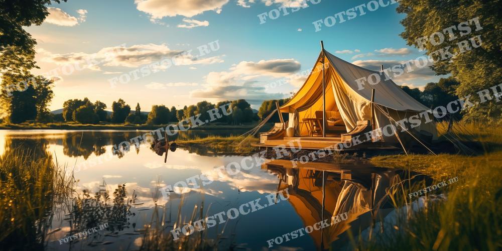 На фотографии изображен глэмпинг на берегу озера в летний вечер. Просторная палатка установлена на деревянной платформе, окруженной зеленью и деревьями. Озеро спокойно отражает небо, палатка и природа вокруг. Внутри палатки можно увидеть уютные детали интерьера, создающие атмосферу комфорта и релаксации. Это изображение может быть использовано для рекламы глэмпинг-туров, туристических агентств, а также в статьях и блогах о природе и отдыхе на свежем воздухе.