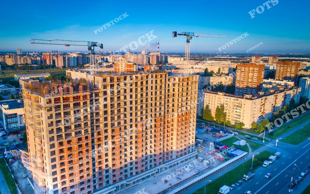 На фотографии изображен процесс строительства крупного жилого дома в городской среде. Подъемные краны и строительные леса подчеркивают масштаб проекта. Снимок символизирует рост города, развитие инфраструктуры и современные методы строительства.