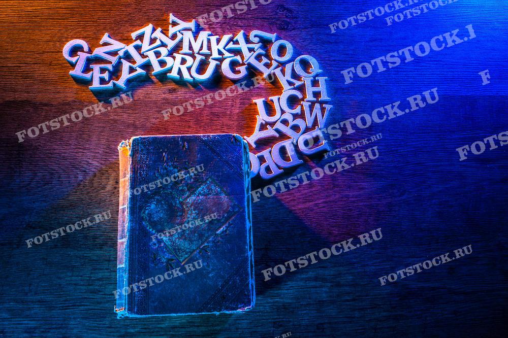 Фотография показывает старинную книгу и разбросанные белые буквы, лежащие на деревянном столе. Эта сцена символизирует распространение знаний и литературы.
