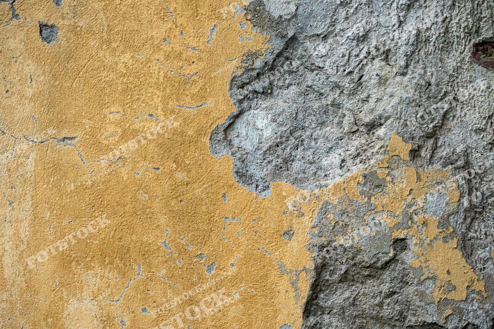 Изображение стены с облупившейся желтой штукатуркой, оголяющей грубую текстуру серого камня. Контраст между яркой краской и изношенной поверхностью подчеркивает воздействие времени и атмосферных условий.
