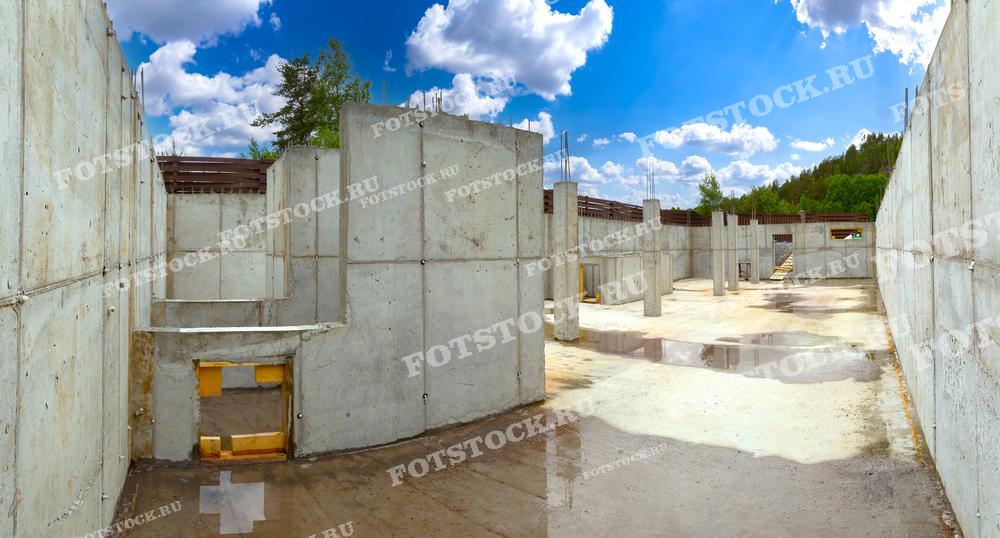 На фотографии изображен фундамент и бетонные стены на строительной площадке, находящиеся на стадии возведения. Бетонные конструкции поднимаются вверх, образуя основу для будущего здания. На полу видны лужи, отражающие ясное голубое небо с облаками. Окружающая зелень и забор на заднем плане подчеркивают естественное окружение и текущий этап строительства. Фотография иллюстрирует важность надежного фундамента и строительных работ.