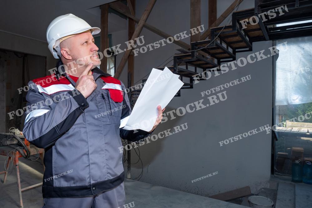 На фотографии изображен инженер в защитной каске и рабочей форме, держащий в руках планы и документы. Он находится на строительной площадке, внимательно изучая проект и оценивая ход строительных работ. Фото иллюстрирует процесс контроля и проверки строительства, демонстрируя профессионализм и ответственность специалиста.