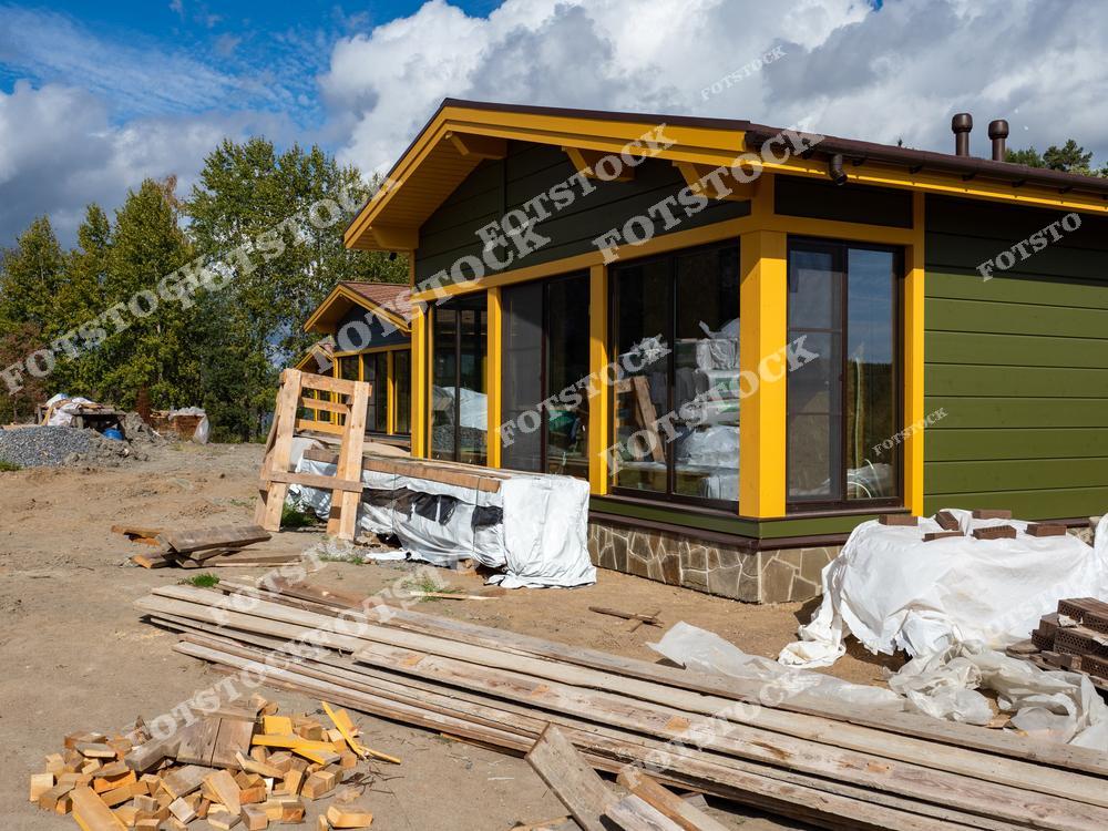 На фотографии изображен недостроенный дом с большими окнами и ярко-желтой отделкой, окруженный строительными материалами и инструментами. Территория вокруг дома усеяна досками, кирпичами и упаковками. Снимок подчеркивает процесс загородного строительства на финальных стадиях